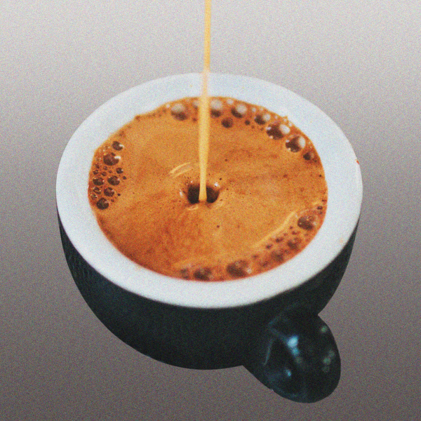 A small cup being filled to the brim with espresso, on a dark grey gradient background.